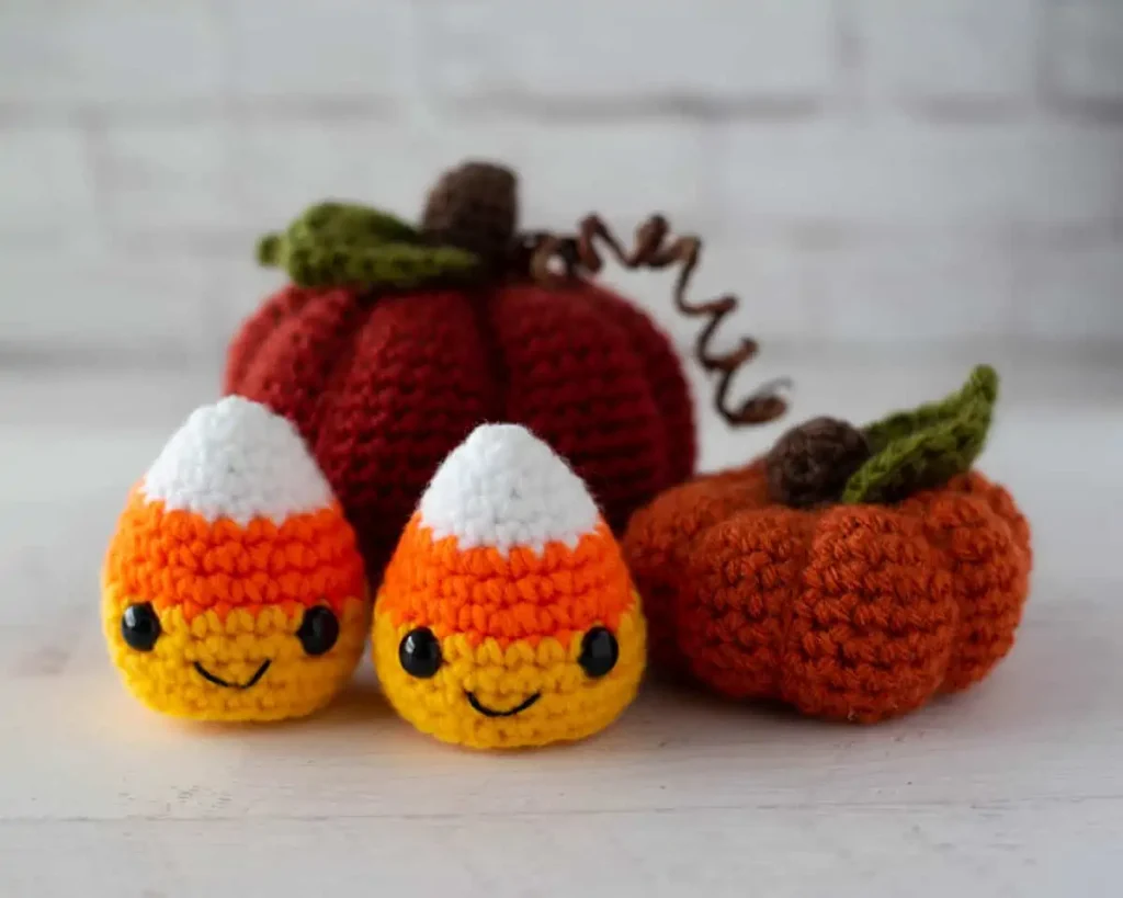 Two crocheted candy corn figures with smiling faces sit in front of two crocheted pumpkins, all arranged on a white surface with a white brick wall background.