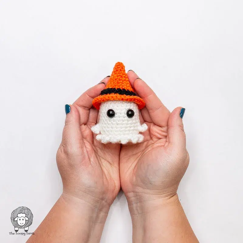 Two hands hold a small crocheted white ghost with black eyes, wearing an orange and black witch's hat, against a white background.