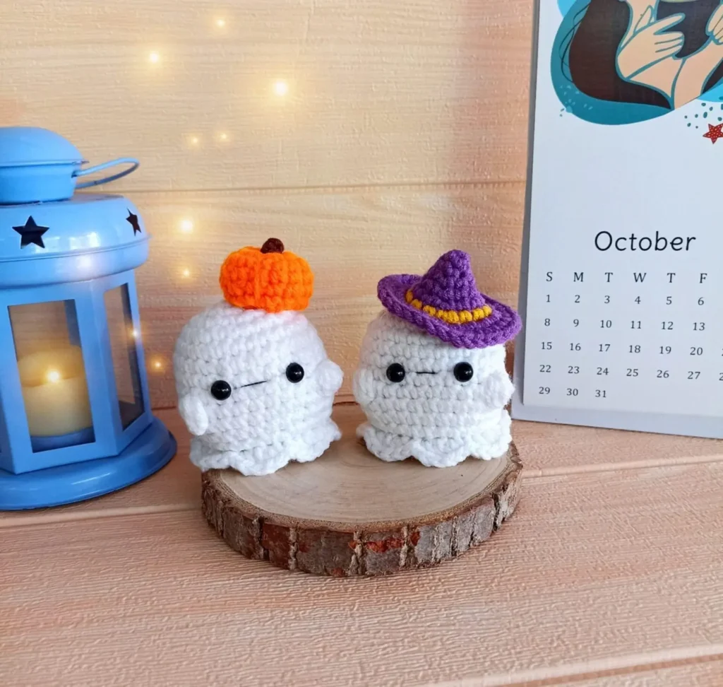 Two crocheted ghost figures, one with a pumpkin hat and one with a purple witch's hat, sit on a wooden slice next to a blue lantern and an October calendar.