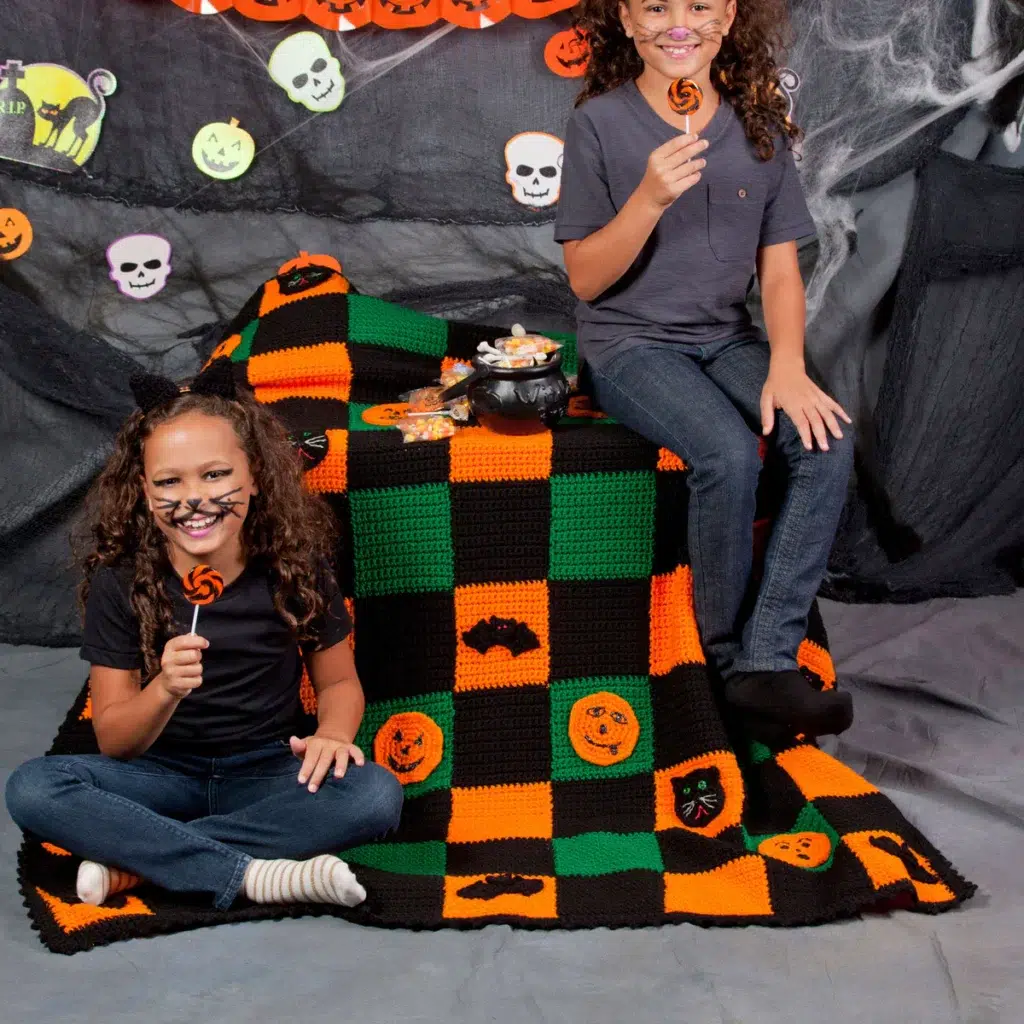 Two children in casual clothes with face paint sit on and next to a Hallowe'en-themed blanket, holding orange lollies, surrounded by spooky decorations, treats, and playful creations inspired by free Hallowe'en crochet patterns in the background.