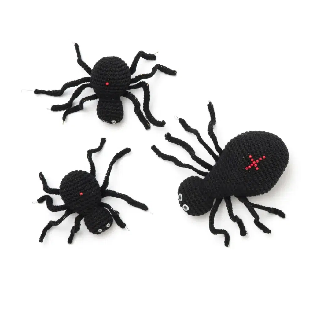 Three crocheted black spider figures with red yarn markings and googly eyes are arranged on a white background, making them the perfect addition to your collection of free Hallowe'en crochet patterns.