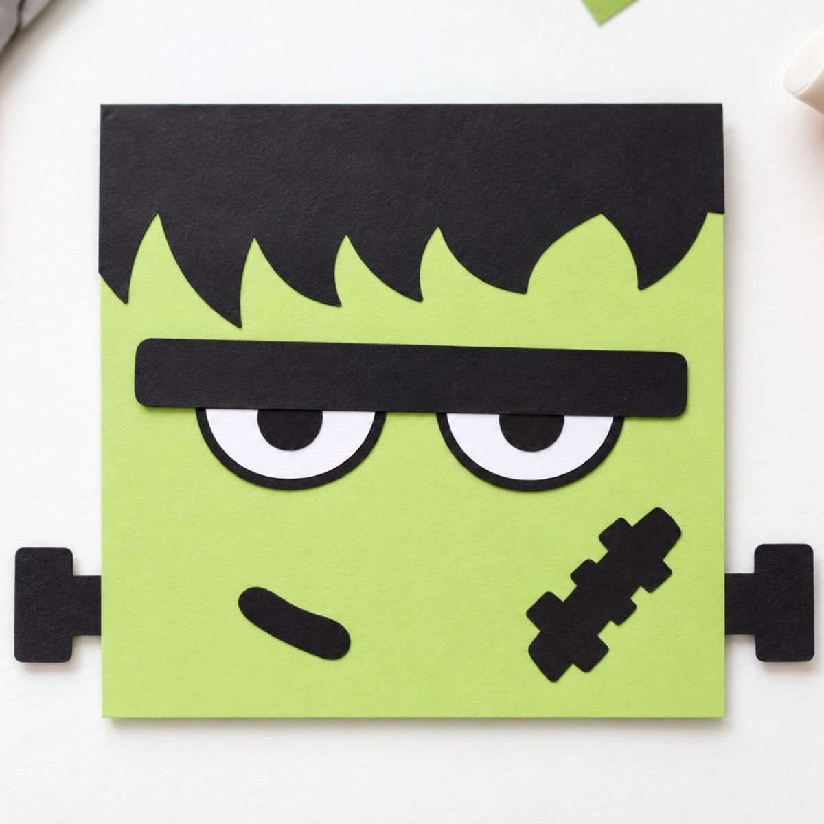 Square paper craft of a green Frankenstein face with black hair, eyebrow, bolts on sides, and a stitched scar on the cheek.